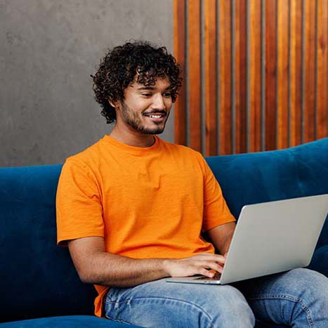 Young IT Indian man wear orange casual clothes sit on blue sofa couch hold use work on laptop pc computer stay at home hotel flat rest relax spend free spare time in living room indoor Lounge concept